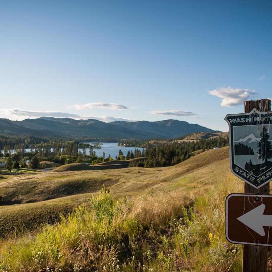 A Washington State Parks logo sign points towards Lake Curlew that is visible beyond golde, grassy, rolling hills.