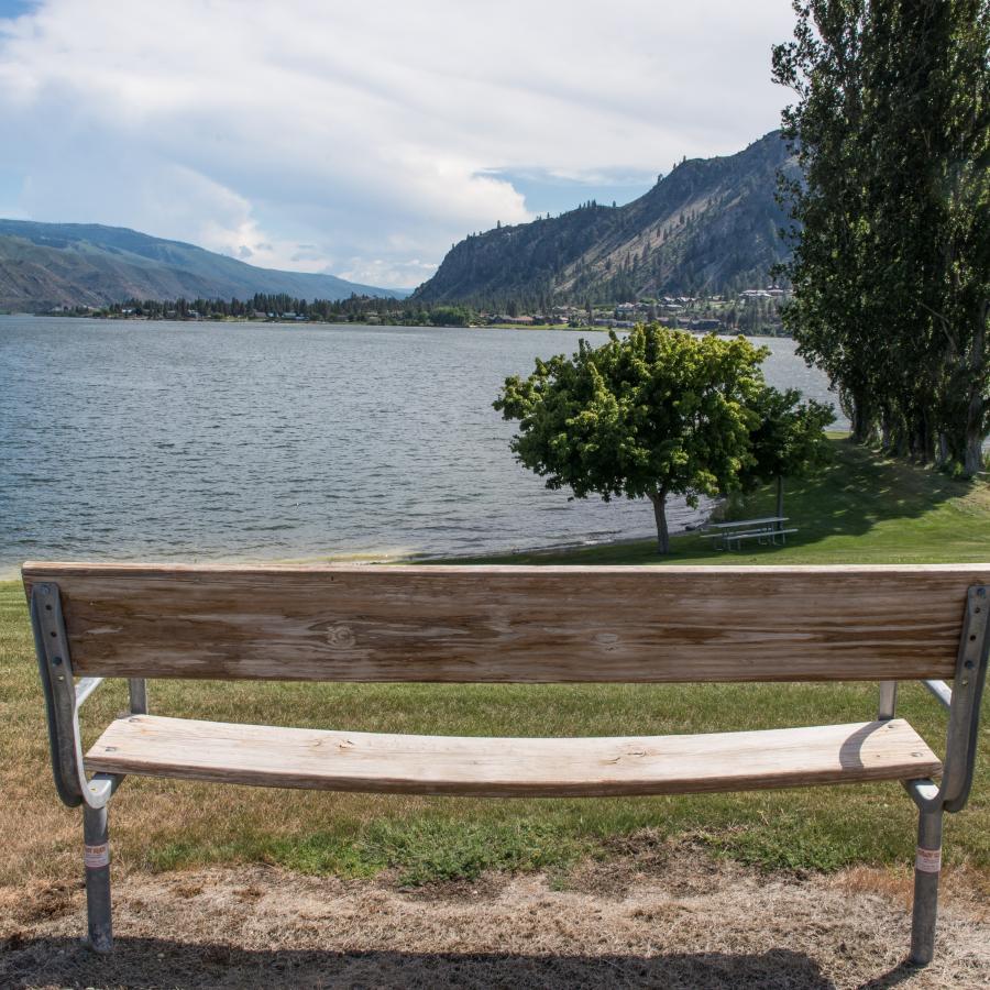 A wooden bench seen from behind sits not far from the river's edge with tall mountains in the background