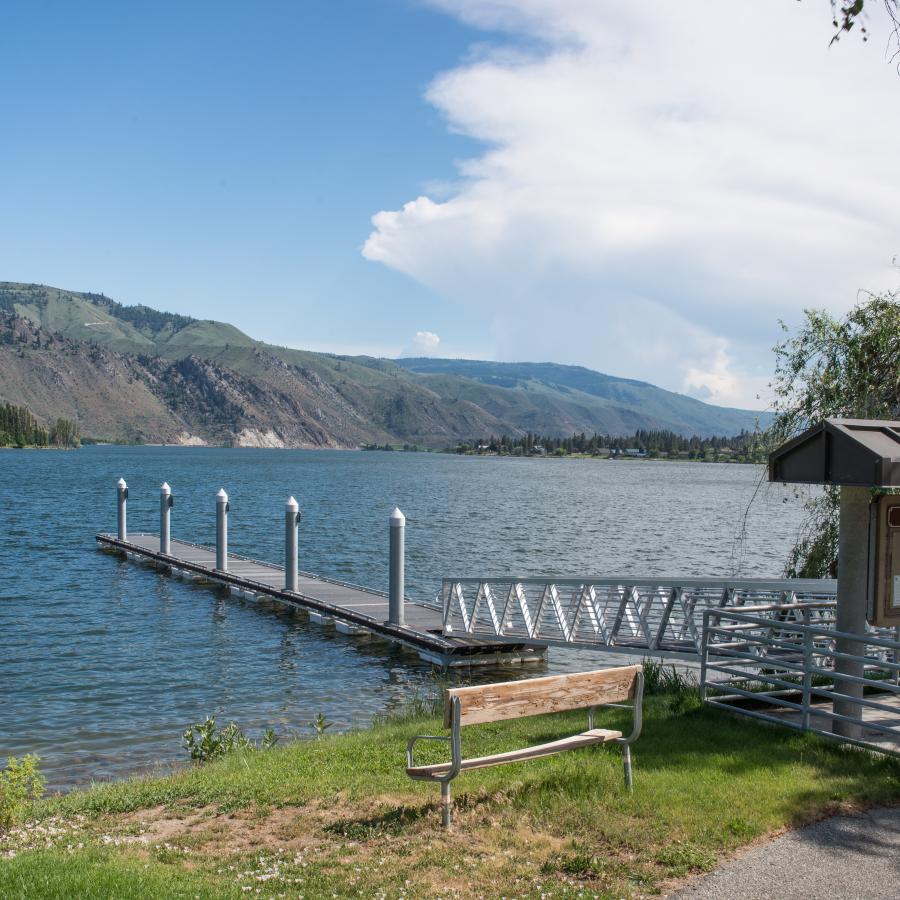 A metal ramp leads down to a dock at the edge of the river with a wooden bench nearby and tall mountains on the opposite side of the water.