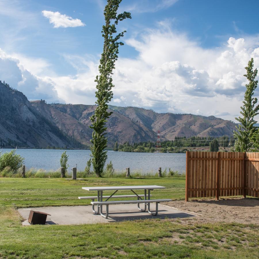 A picnic table sits on a concrete pad. Lawn extends to the edge of the river and tall mountains stick up prominently along the far side of the water.