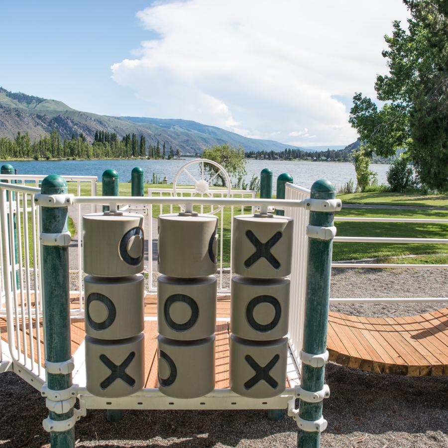 A children's playground with a green lawn and pathways leading down to the water. Tall hills and trees are in the background.