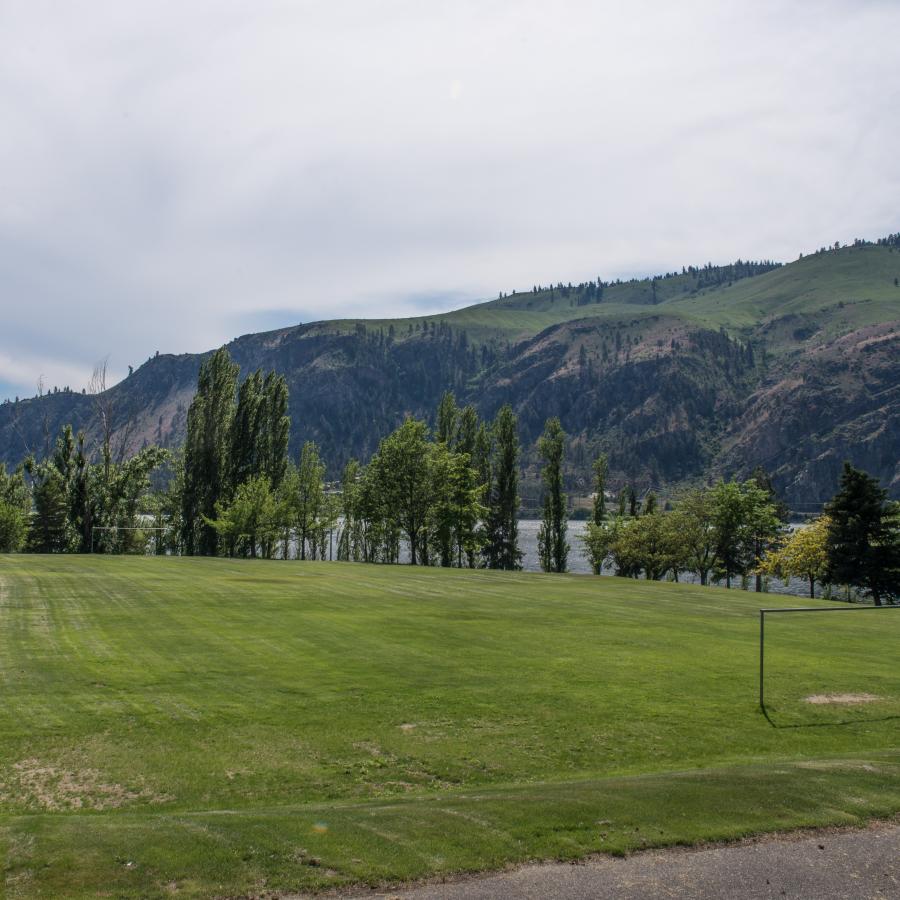 A large grassy sports field with a view of the river through trees. Tall mountains are along the far side of the water.