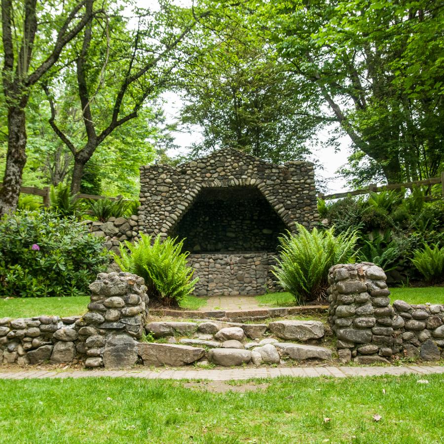 The grotto is made up of hundreds of stones of varying colors surrounded by multiple bushes and lush green trees. There is a path leading up to the grotto with a few steps.