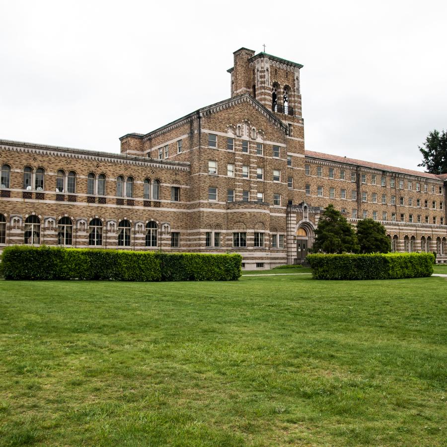 The Seminary at Saint Edward State Park where the Saint Edward Lodge can be found. The building is very long with a center building that appears to be 2 floors higher than the rest of the building. There is also a bell tower that reaches above the tallest part of the building. The lawn area of the seminary is also visible with bright green grass and well manicured hedges,