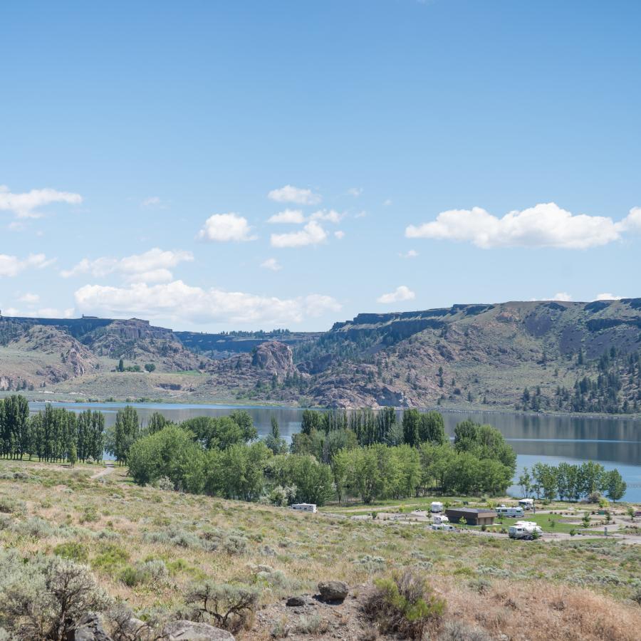 View of campground with green trees and the lake behind, reflecting the basalt rock hillside in the background