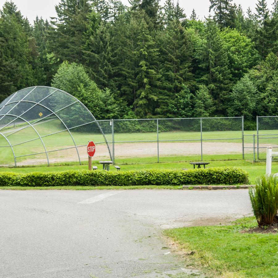 Ballfield with a fence on two sides and the other two sides bordered by forest. The fence runs along the road and a stop sign is visible.