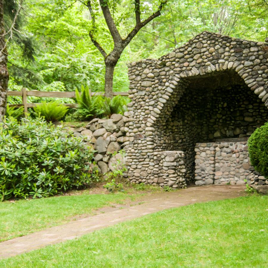 The stone grotto is made up of hundreds of stones of different colors including reddish brown, tan, brown, grey-brown, grey, and shades in between. The grass is green and a path leads up the center to the grotto. The plants visible around the grotto are green with one bush on the left displaying a single purple flower.