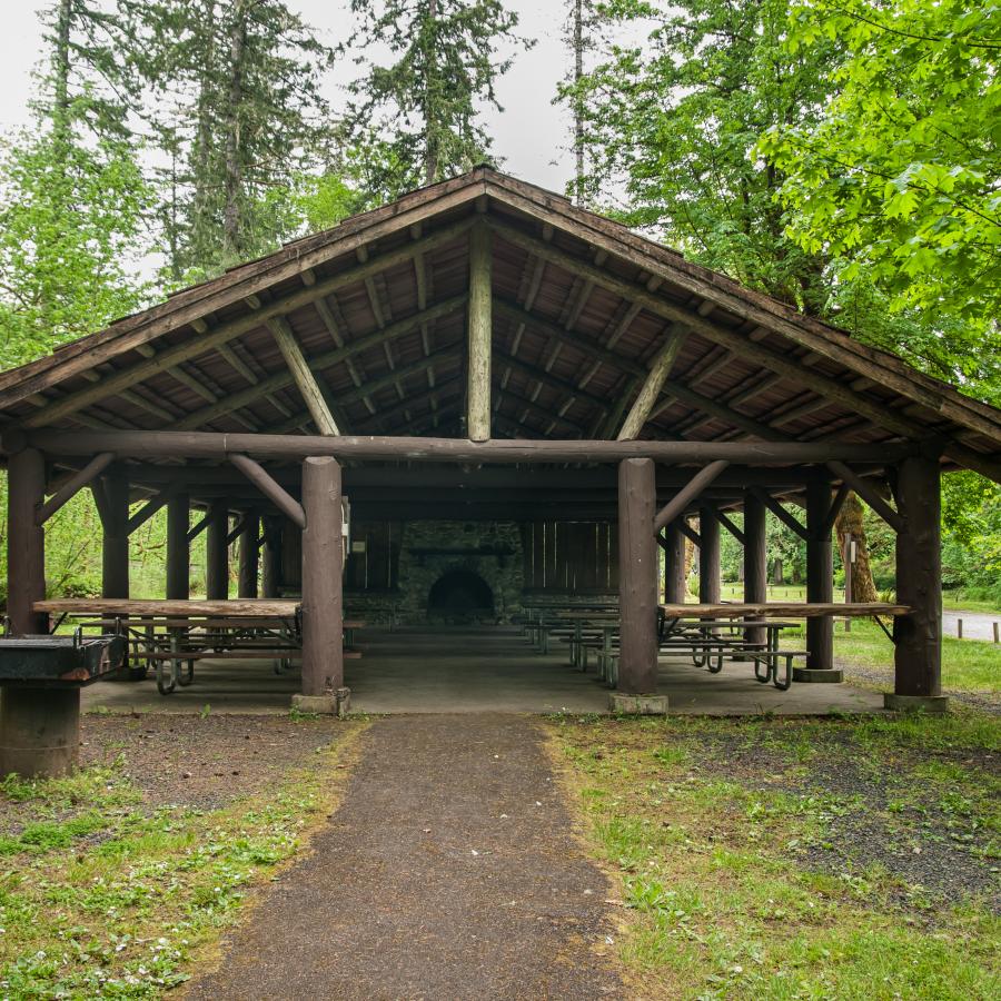 Kitchen Shelter with large wood log columns, a stone fireplace, a wooden roof, and many picnic tables. There is a large grill and a path leading to the shelter. 