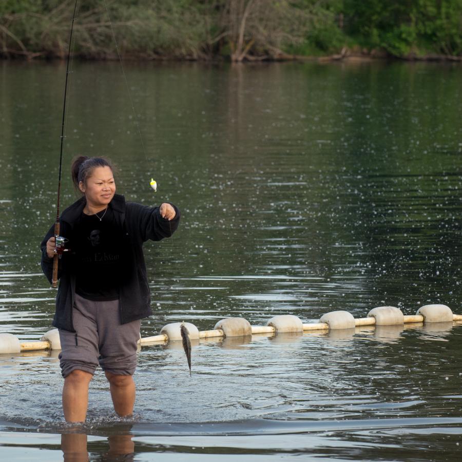 A feminine presenting person dressed in a black fleece and black t-shirt with pants pulled up to their knees is fishing in the waters of Lake Sammamish. The person is standing in shin-deep water and has caught a small fish on their fishing line. 