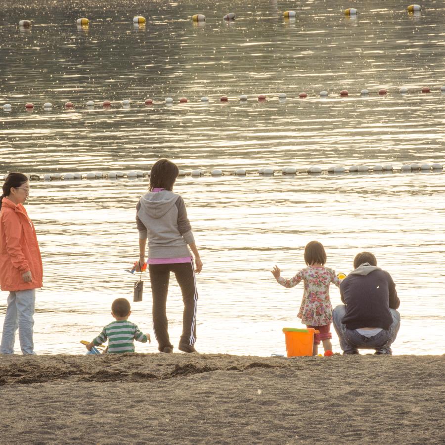 Family with 2 young children and 3 adults exploring the sand at Sunset Beach. One adult is wearing light colored jeans and a peach colored jacket, another is in black track pants and a grey and pink sweatshirt, and the third in a grey or blue sweatshirt and jeans. One of the youth is wearing a green and white stripped shirt and the other is in a floral pattern. The swimming buoys are visible on the water and it appears to be golden hour where the sun creates a soft, golden glow. 