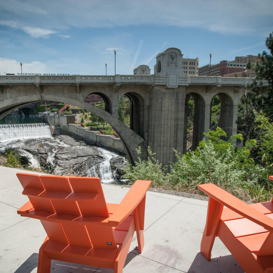Two chairs sit along the edge of the Centennial Trail. A waterfall is visible underneath a large concrete bridge.