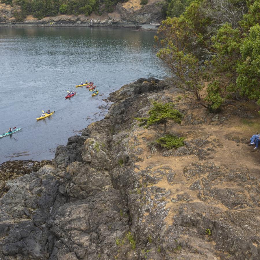 A group of six kayaks paddle along the shoreline of the rocky cliff as two hikers take a rest at a picnic table to watch the kayakers pass by. There are green trees and shrubs  around the onlookers.
