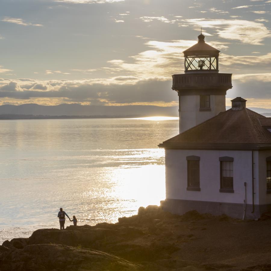 A small lighthouse, sitting on a rocky cliff, with the sun setting behind. An adult holds the hand of a child as they walk along the rocks to the left of the lighthouse.