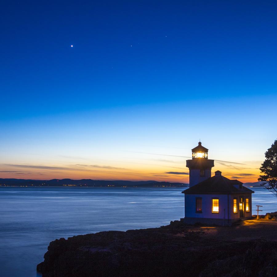 An illuminated lighthouse sits on a rocky cliff overlooking water as the sun sets behind and the sky grows dark. A star shines bright in the sky.