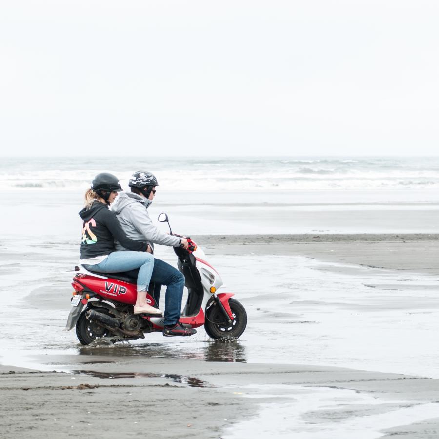 two people riding red scooter on hardpacked sand of the beach by shoreline through receding  pools of water