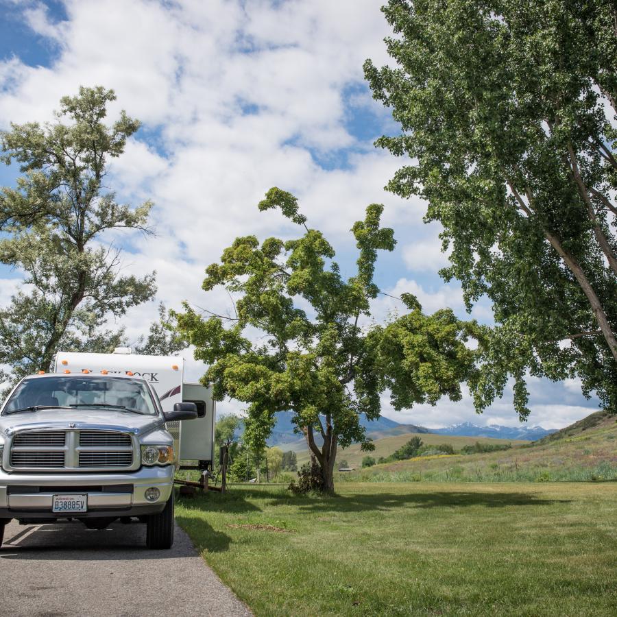 Big silver truck sits in front of a camper trailer at a campsite, surrounded by grass and trees; hills and mountains can be seen in the distance behind the campsite.