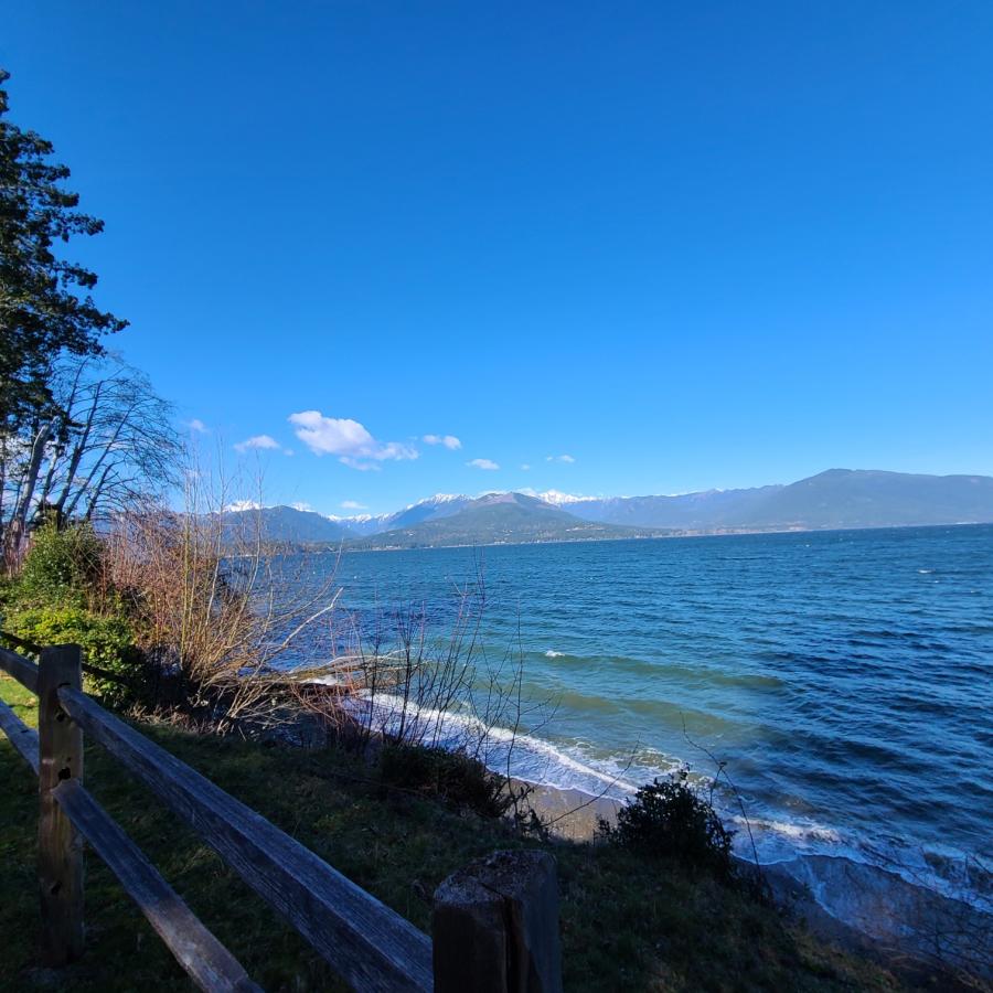 The shoreline at Scenic Beach State Park, with a rocky shore and green leafy trees in the foreground and brilliant blue water in the background. In the distance, the Olympic Mountain range rises into a clear blue sky.