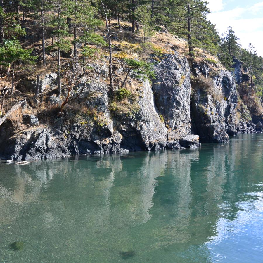 View of rugged cliffs along the James Island shoreline on a sunny day. The water is clear and you can see the pebbly floor beneath it.