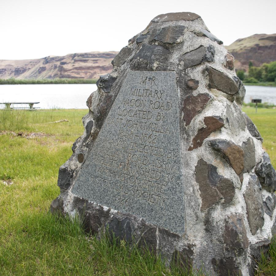 A rock monument, dedicated in 1926 to memorialize the Military wagon road in 1858-62, sits in the grassy lawn. The river and hillside can be seen in the background.