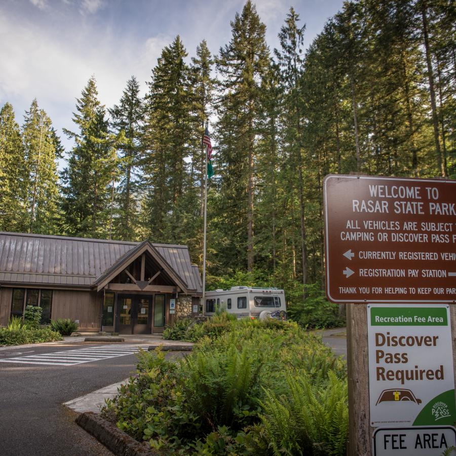 Image of the Rasar visitor center in the center of the photo with wooden, brown siding and a brown roof. There is an RV visible to the right of the visitor center, a flag poll with the American and Washington State flags, and greenery visible in the foreground. There is a sign in the foreground welcoming you to Rasar state park with directions on how to register and notifying visitors that a discover pass is required. 