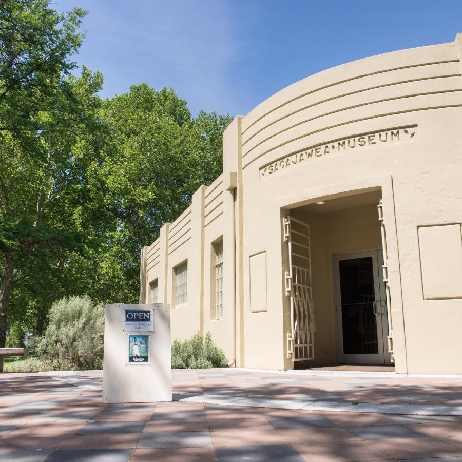The tan interpretive center is surrounded by large green, shade trees, water and green grass can be seen in the distance. A bench sits outside the center on red and grey concrete pavers with an open A-frame sitting in the middle of the cement pavers.