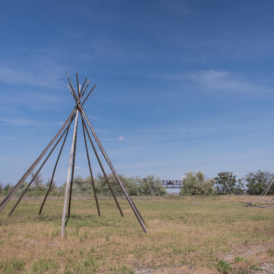 Two uncovered teepees are set in a brown and green field with blue skies and wispy clouds. Trees line the background with a bridge poking out between them.