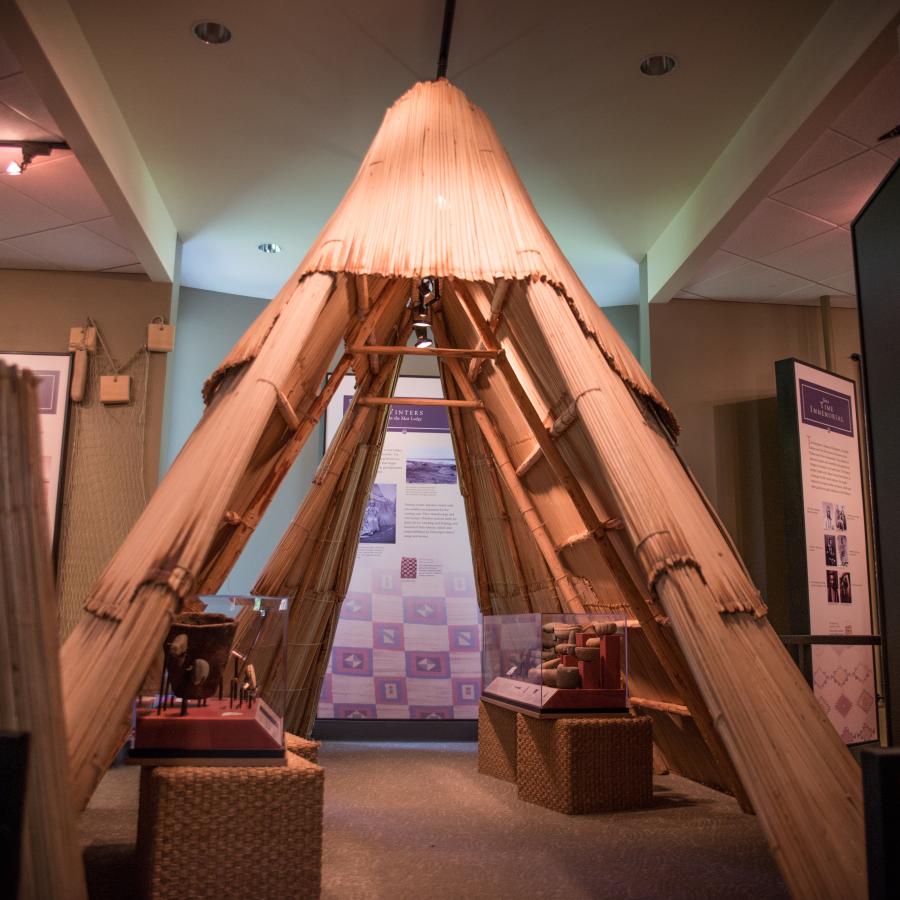 Inside the Interpretive Center, a replica of a teepee sits in the middle of the room. Tools and a bowl are on display under the teepee. A quilt display can be seen behind the teepee.