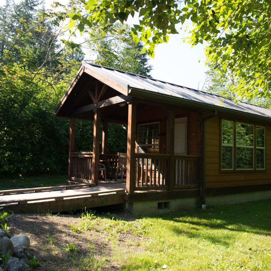 Front and right side view of a reservable cabin available at Wallace Falls. There area large windows on the front and side of the cabin as well as a front door that is white. There are a couple chairs on the porch of the cabin which has a boardwalk style trail to the entrance. The cabin is surrounded by lush green forest. 