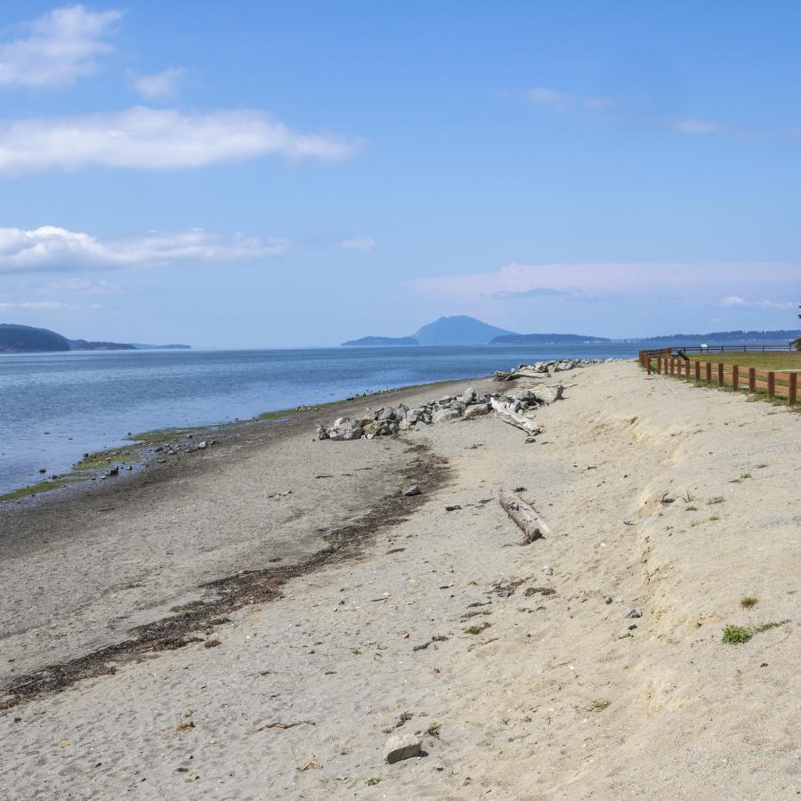 Looking down the beach, lined with drift wood, sits below a grassy field lined with a wooden fence. Blue skies, high water and islands sit in the background. 