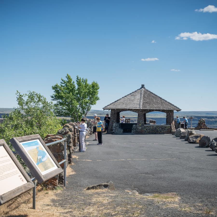 Looking at the rock shelter with a shake roof, with trees and interpretive panels lining the walkway. A blue sky set in the background.