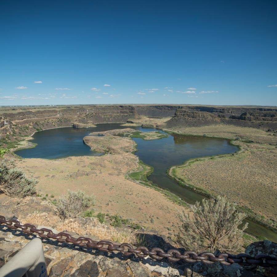 Looking over a rock wall with a chain rope, sits a lake with brown grasses and green shrubs at the bottom of Dry Falls. A blue sky sits in the background
