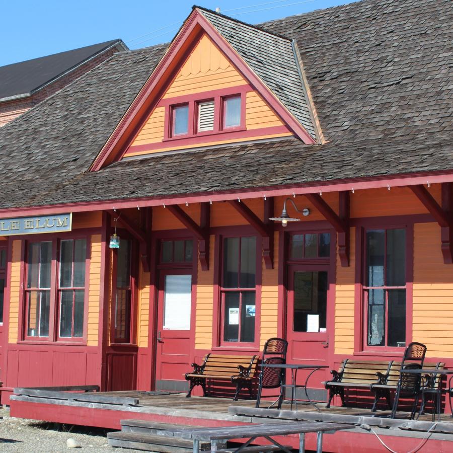 Old western style building painted orange with red trim with a sign on the left front reading, "Cle Elum". Wooden park benches sit on the front, open porch of the building. Picnic tables sit on the dirt in front of the building.