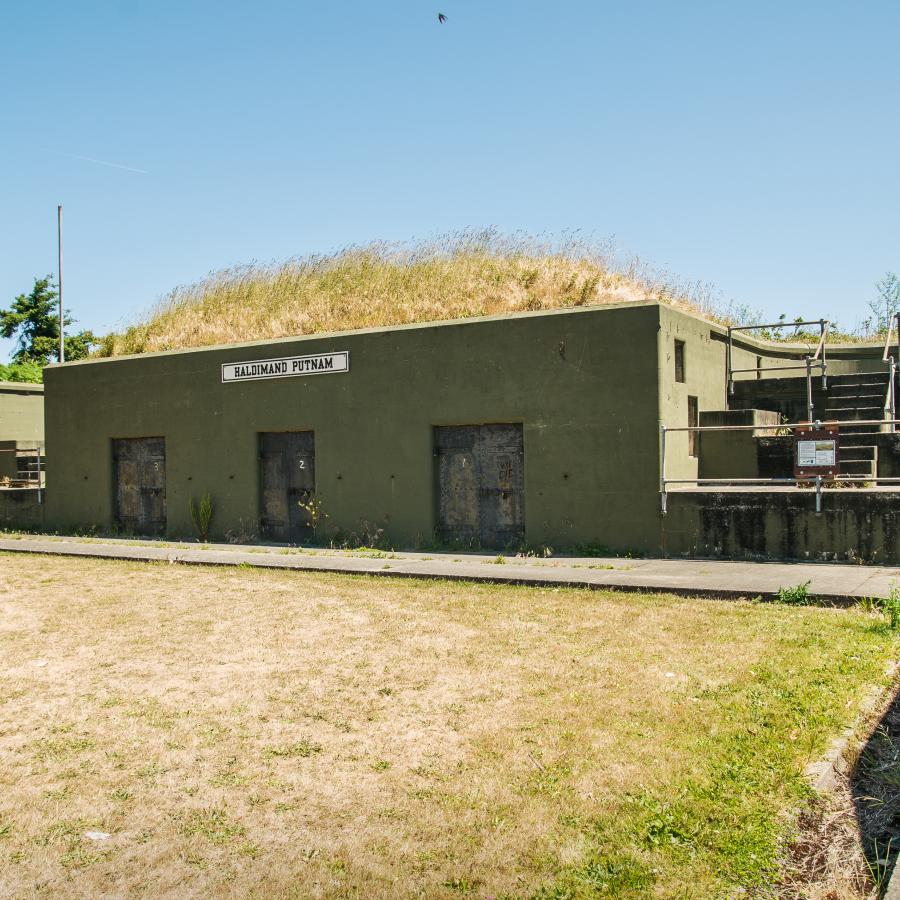 A cement battery building sits with brown grass in front and on top. Doors are boarded shut with stair ways throughout the structure.