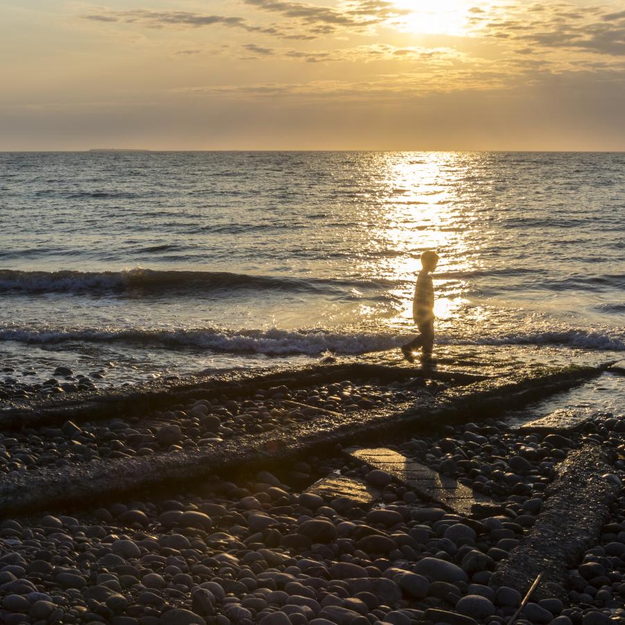 A child walks along the waters edge at sunset with the waves rolls in and a pale orangish cloudy sky in the background.