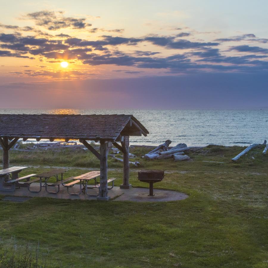 A wooden picnic shelter with picnic tables, surrounded by green grass, sits near the beach with driftwood nearby. The waters of the Strait of Juan de Fuca with a blue, yellow and pink sky as the sun sets behind the shelter.