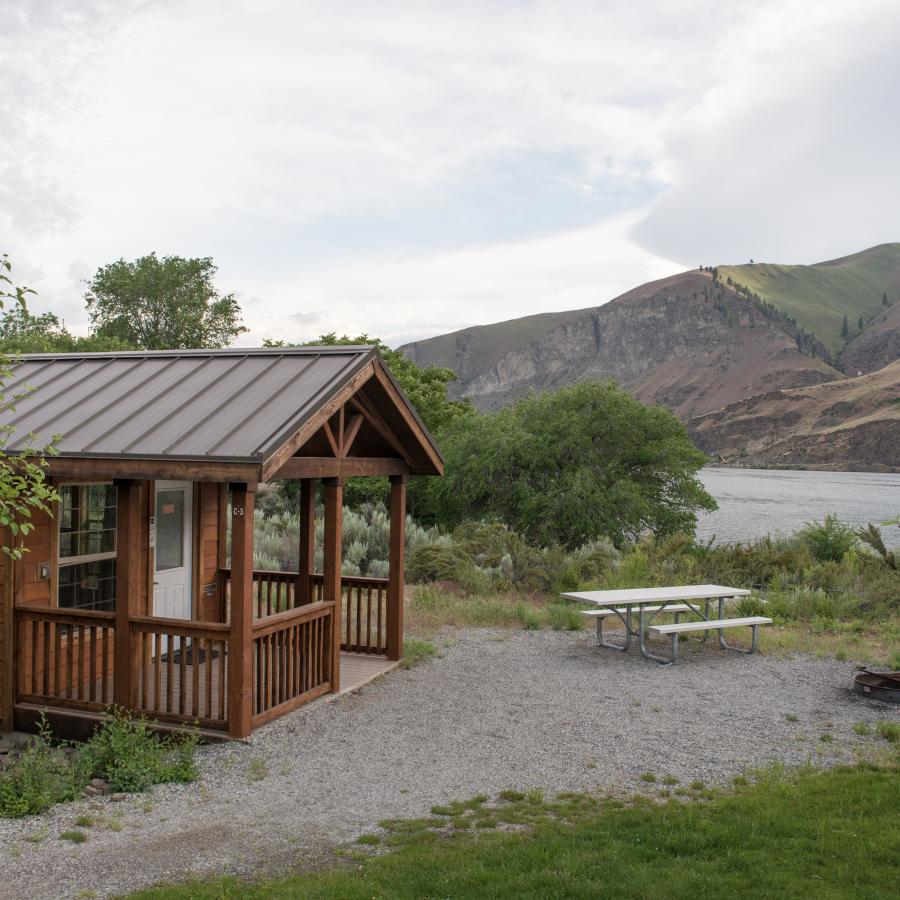 A wood cabin sits in front of the water with a gravel pathway leading to the front door. A picnic table and firepit sit in front of the cabin. Green grass and bushes surround the area. Green and brown hillside with a cloudy sky sit in the background. 