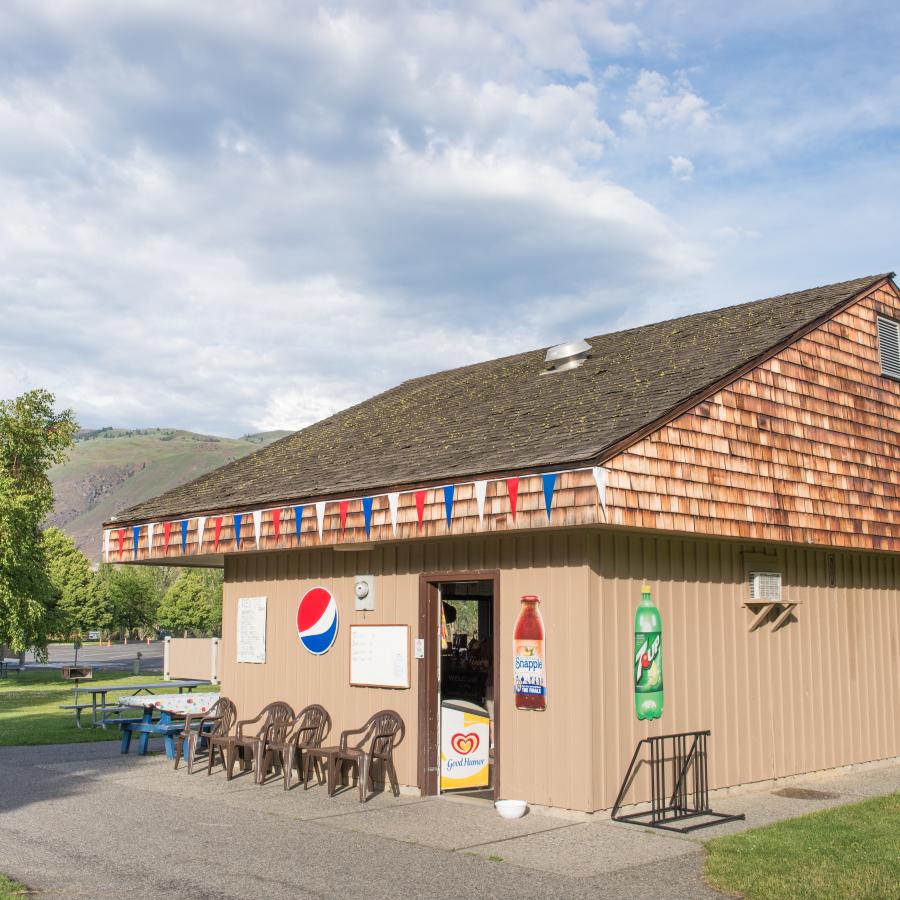 A tan concession building with red, white and blue flags hanging on the fascia and chairs lining the building. Grass surrounds the building with trees in the lawn. Green and brown hills are in the background with a cloudy ski. 