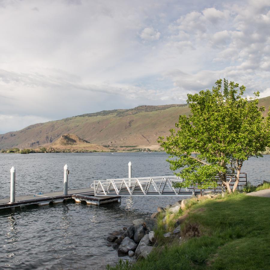 A side view of a T-shaped dock with a single tree at the top of the dock ramp. Brown hills can be seen in the background with a cloudy sky overhead.