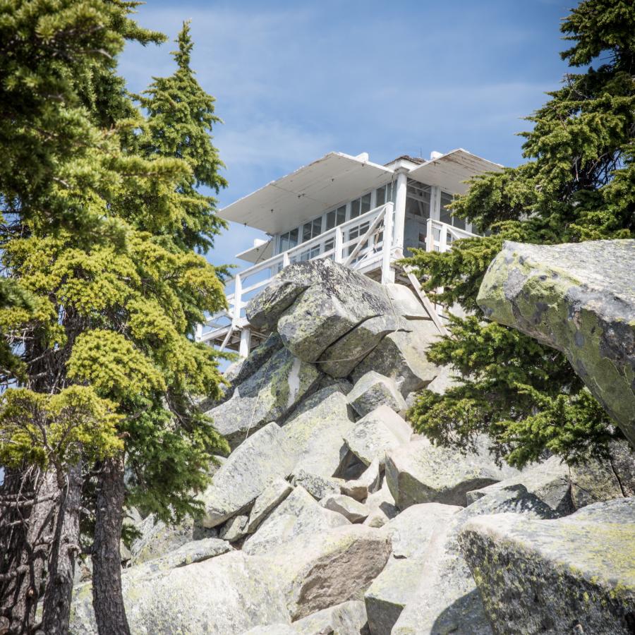 Looking up a rocky terrain with evergreen trees on the sides, at the white lookout tower with the shutters propped open. A blue sky set in the background. 