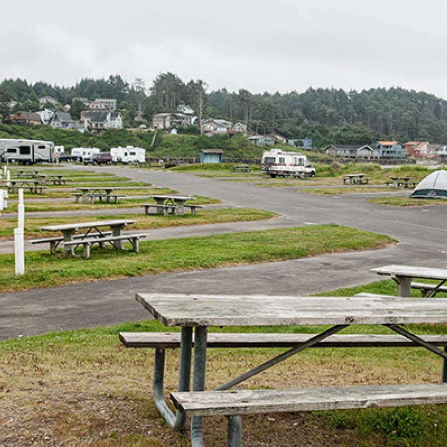 Picnic tables located at RV camping spots next to the Pacific Ocean shore. In the distance are houses and tree-covered sea cliffs