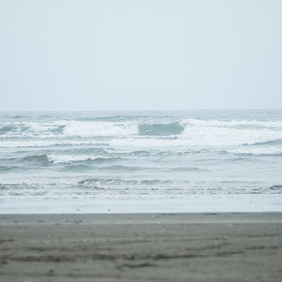 A flat, sandy beach meeting with rolling Pacific Ocean waves under a pale blue sky