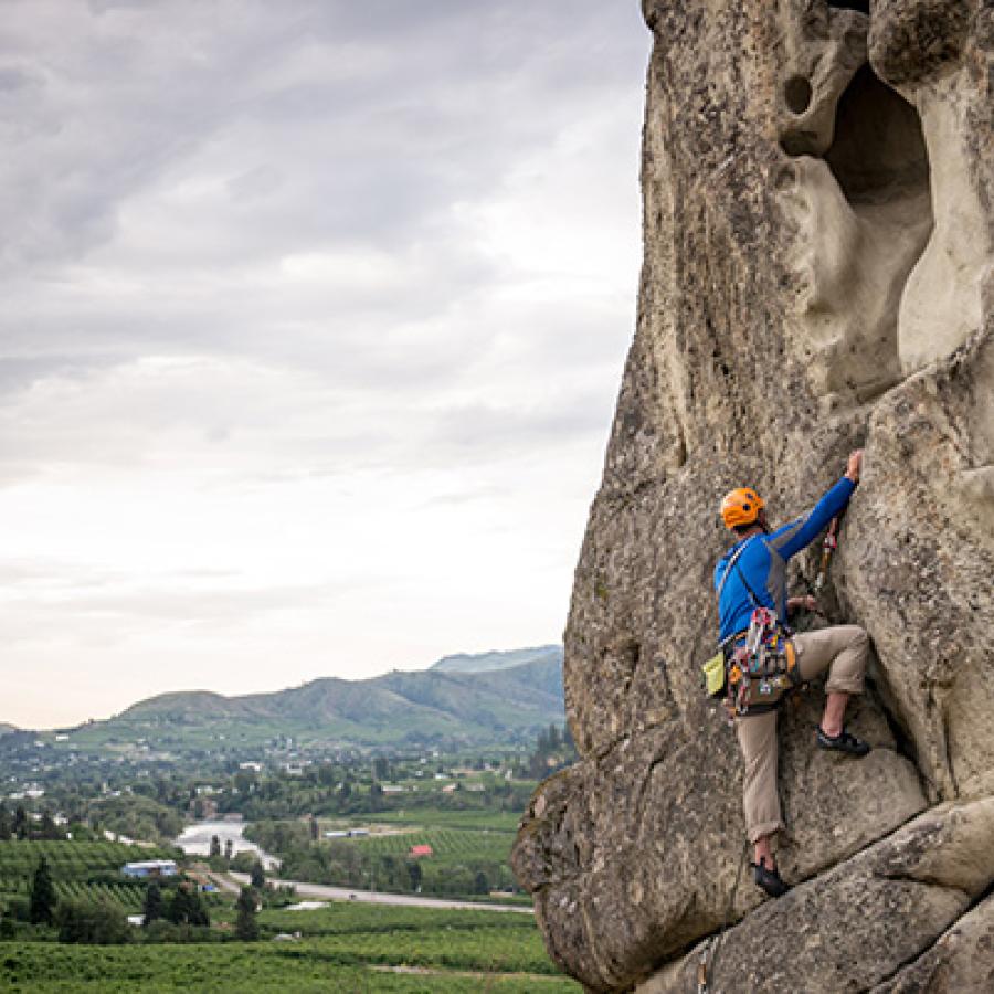 A person in a blue shirt and orange helmet scales a granite rockface using ropes. They look toward the distant verdant valley and mountain ridges under a steely gray cloudy sky.