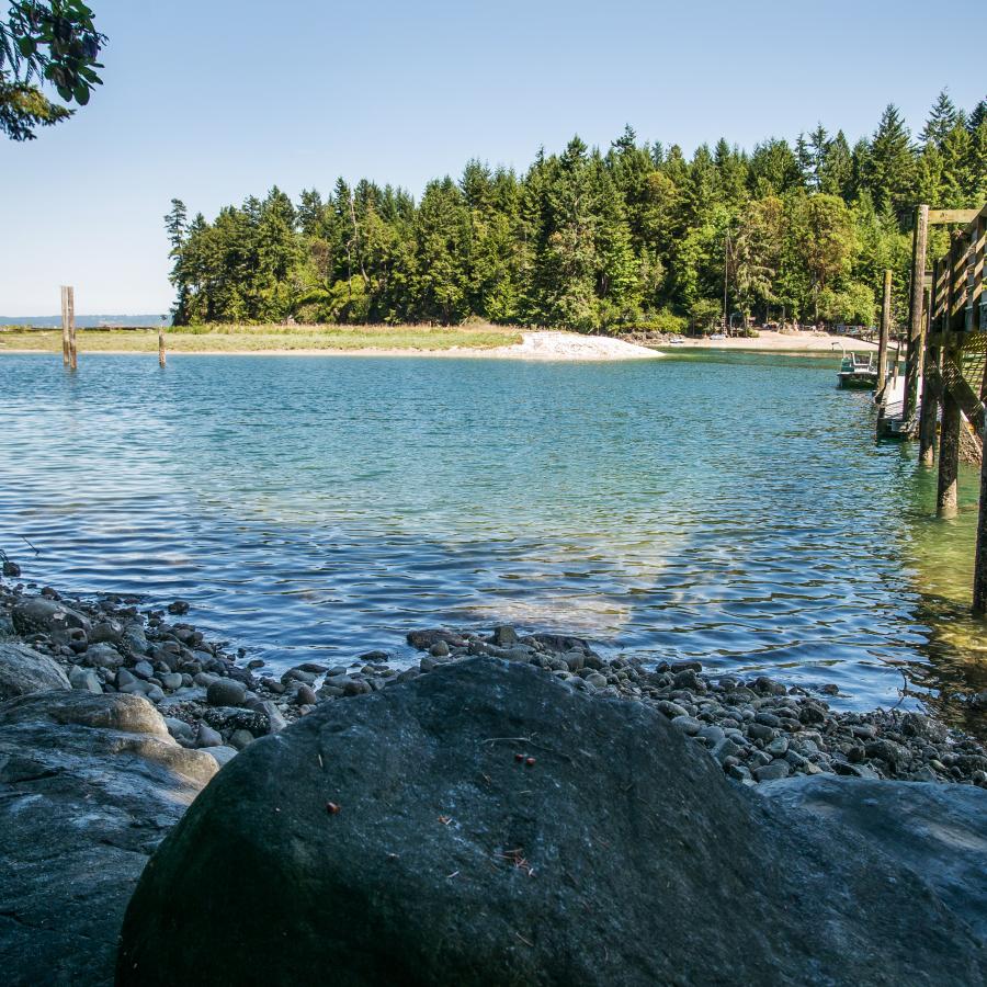 At the rocky water's edge, looking at blue-green water, a  white boat with a green strip sits behind an evergreen tree on the left, across the water are more evergreen trees. A wooden dock sits on the right side with a boat tied to the end of the dock.