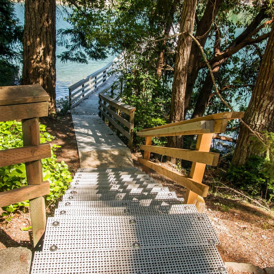 Looking down a set of grey stairs with wood rails, sites the start of the grey dock, tall evergreen trees and blue-green water. A small wood shake roof can be seen behind the staircase rails on the left side.