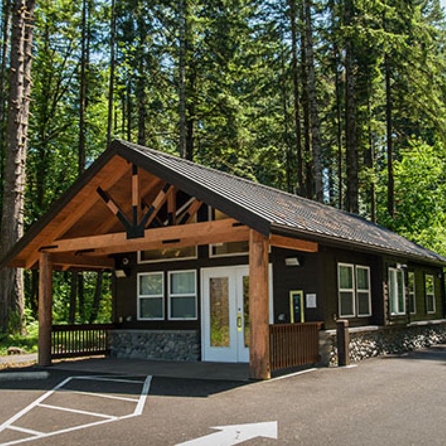 A log-framed ranger station situated on a parking lot. A verdant pine forest rises in the background.