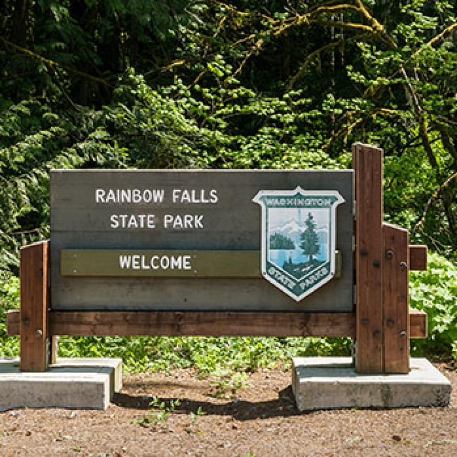 A wood sign reading Rainbow Falls State Park Welcome with the Washington State Parks shield on it sits in front of a lush pine forest.