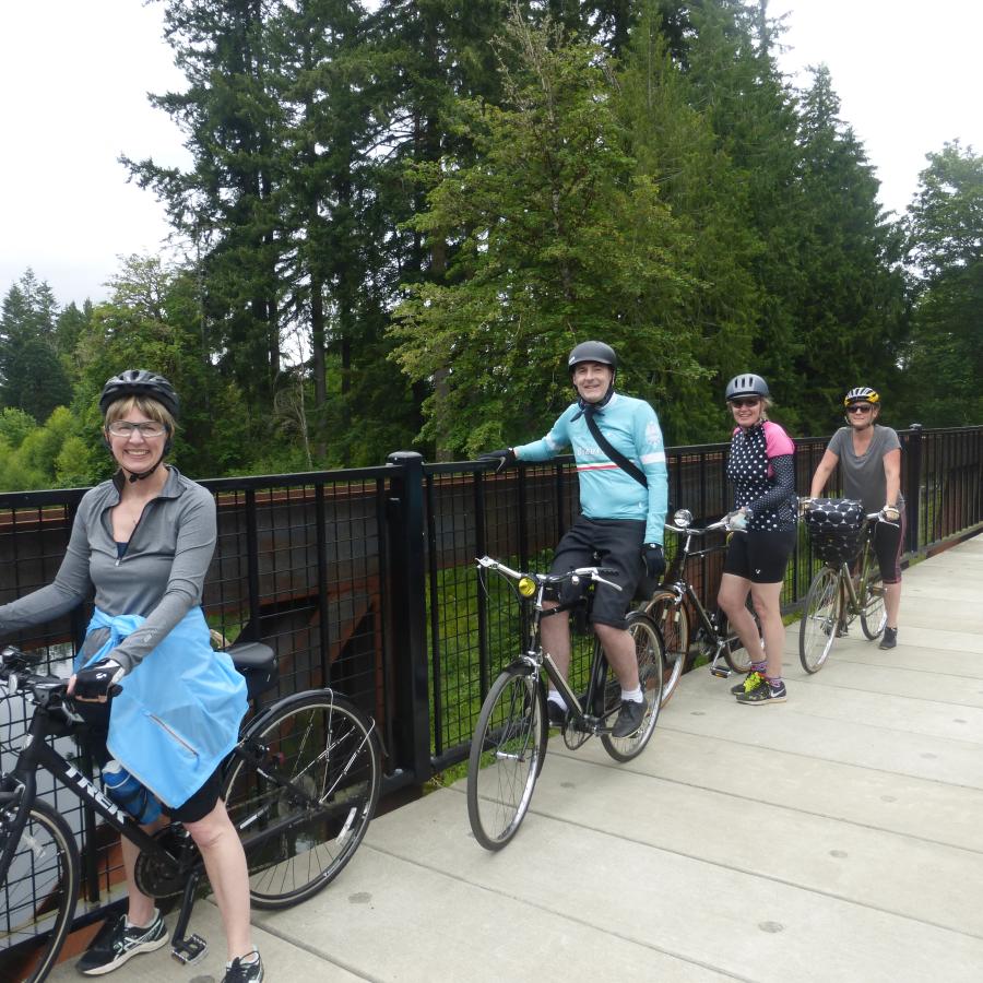 Four smiling people on bikes pose beside the rail of an old railroad bridge that has been converted into a recreational trail.