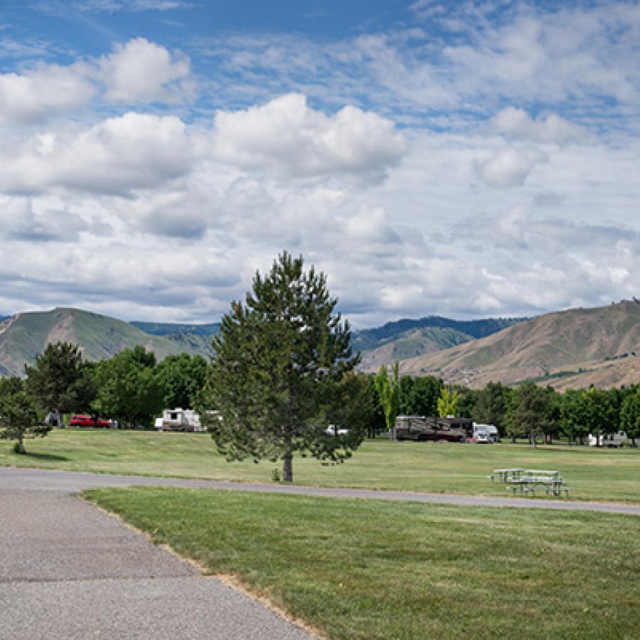 A concrete RV pad in the middle of a wide green lawn under a blue sky full of white, fluffy clouds. High desert hillside rise behind deciduous trees in the distance.