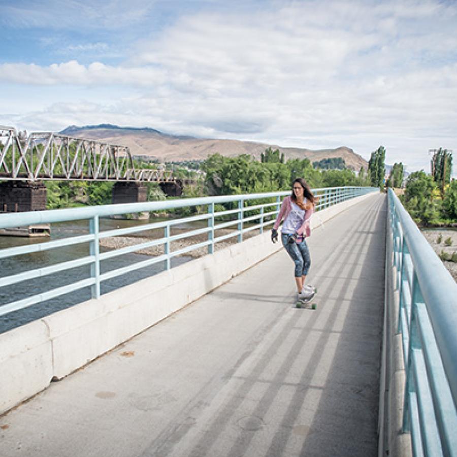 A person cruises over a walking bridge with pale blue railings. To the left a train bridge crosses over the rive to the tree lined shore in the distance.