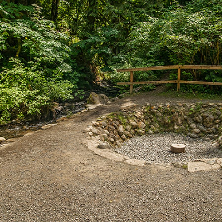 A circular pit surrounded by trees and a wooden fence. It is lined with stones and in the middle is a round stone with a hole in it. This is where the flame of Flaming Geyser was once constantly burning.